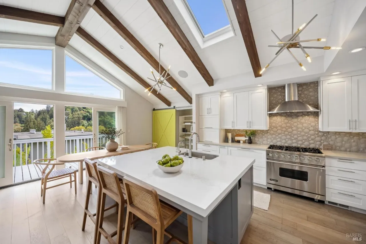 This is a bright and airy kitchen featuring a large island with a white countertop and gray base, complemented by wooden bar stools. The kitchen boasts white cabinetry, stainless steel appliances, and a unique backsplash, with natural light streaming in through large windows and a skylight. Exposed wooden beams add a rustic touch to the modern design.