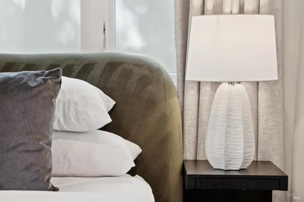 This is a close-up shot of a bedroom featuring a bed with a soft, olive-green headboard. The bed is adorned with white pillows and a gray decorative pillow. A white lamp sits on a dark wood nightstand, adding a touch of elegance to the room. The overall impression is one of comfort and understated luxury.