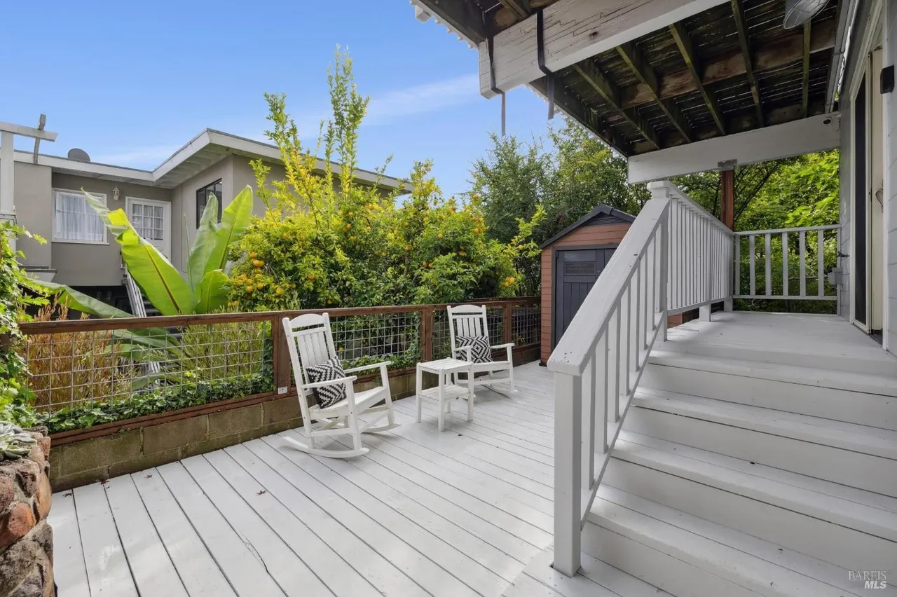 This image showcases a charming outdoor deck area, featuring two white rocking chairs with patterned cushions and a small table. The deck is painted white and is surrounded by a wooden railing with wire mesh, offering a view of lush greenery and a glimpse of a building in the background. A set of white stairs leads up to the deck, adding to the inviting and relaxing atmosphere.