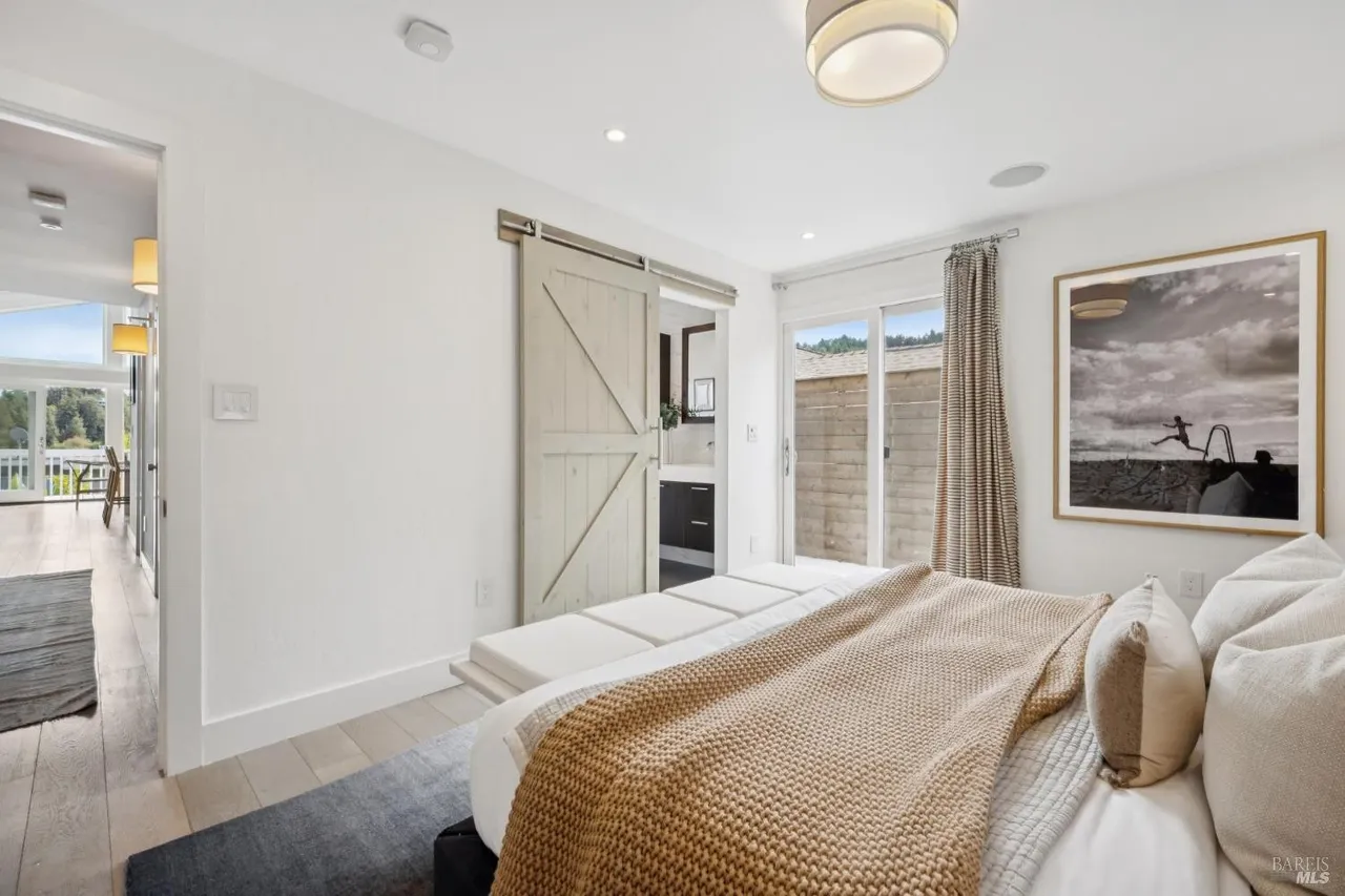 This is a bright and airy primary bedroom featuring a large bed with a tan throw blanket and neutral pillows. A sliding barn door leads to the bathroom, while a sliding glass door provides access to an outdoor area. The room is decorated with a framed art piece and has a modern, minimalist aesthetic.