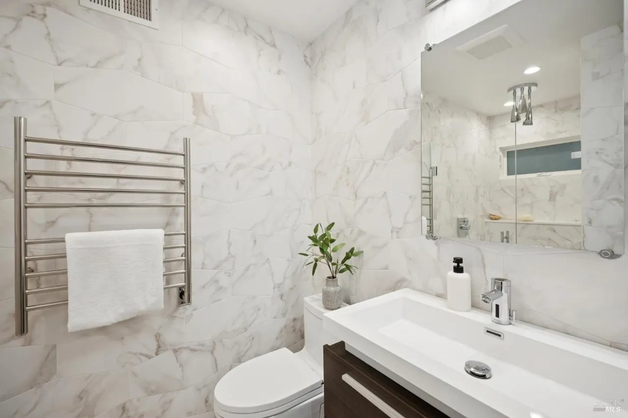 This is a well-lit bathroom featuring white marble-patterned walls and a modern aesthetic. The room includes a sleek white sink with a dark wood vanity, a toilet, and a heated towel rack with a white towel. A mirror reflects the opposite side of the bathroom, enhancing the sense of space.