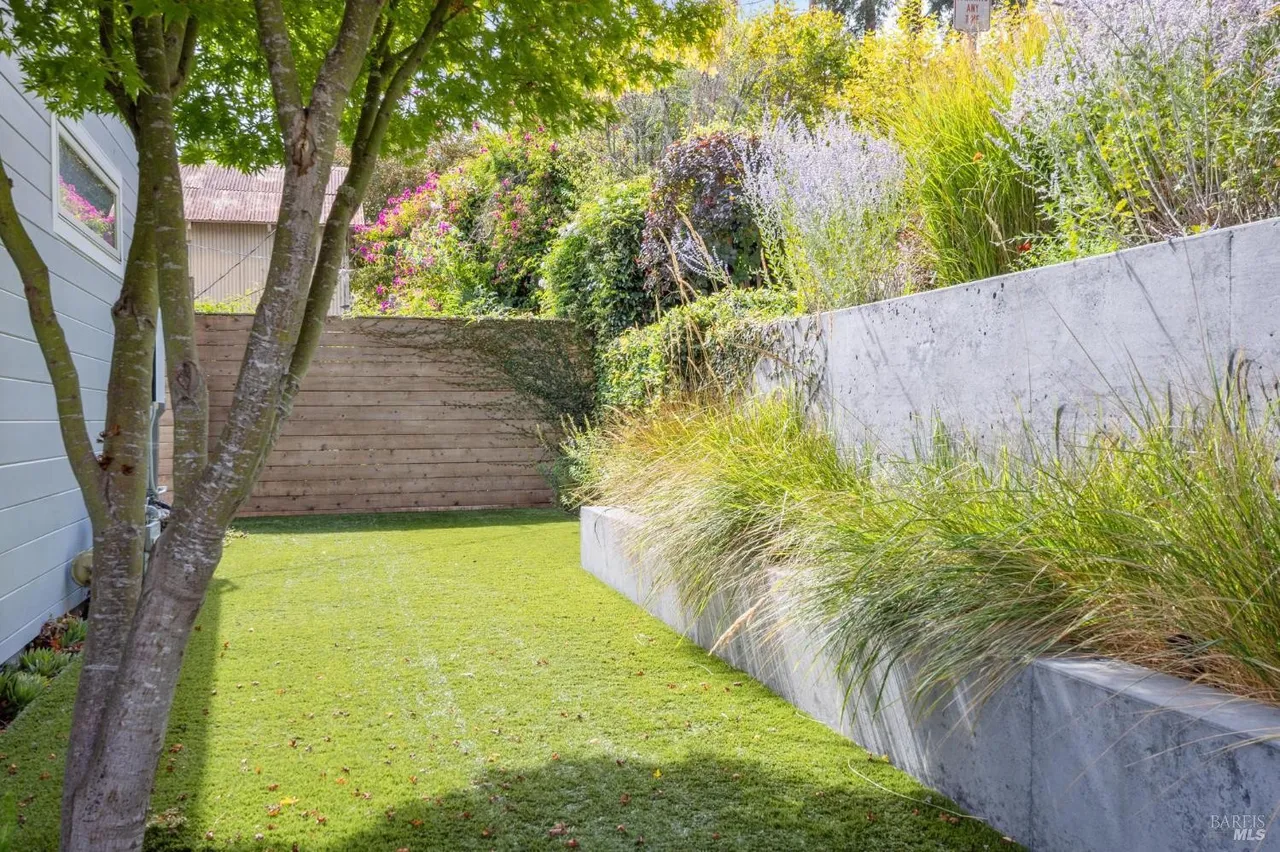 This image showcases a well-maintained backyard garden with artificial turf, a wooden fence, and a concrete retaining wall. Lush greenery and flowering plants add color and texture to the landscape. The yard appears to be a private and serene outdoor space, perfect for relaxation or entertaining.