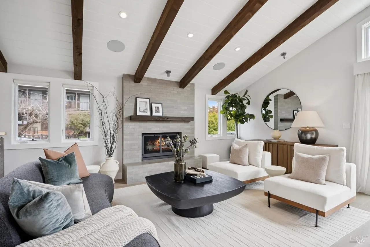 This is a bright and airy living room featuring a modern design with exposed wooden beams and a neutral color palette. The room is furnished with a comfortable gray sofa, two white armchairs, and a unique dark-colored coffee table. A fireplace with a light gray brick surround adds a focal point, and large windows provide ample natural light, creating a welcoming and stylish space.