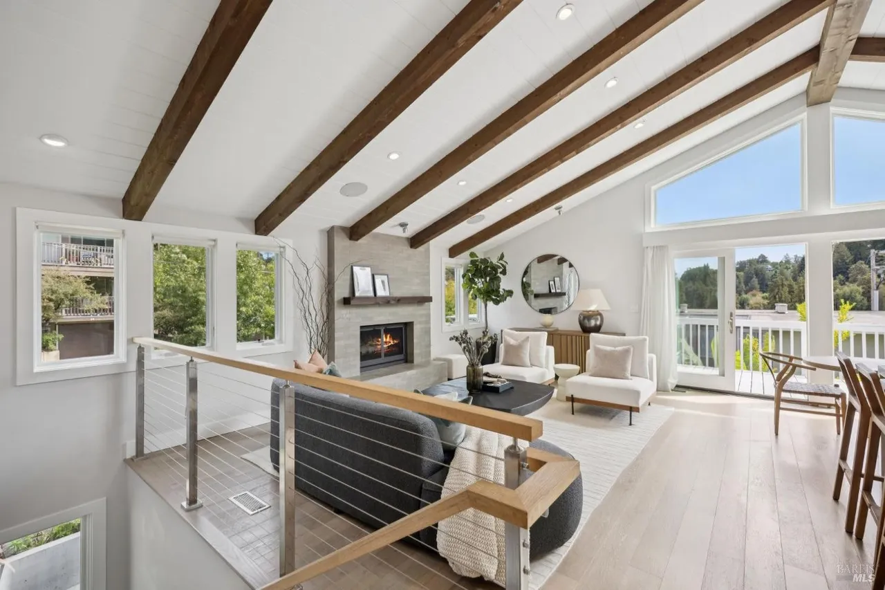 This is an interior shot of a living room featuring a vaulted ceiling with exposed wooden beams, a modern fireplace, and large windows that provide ample natural light. The room is furnished with a gray sofa, white armchairs, and a round coffee table, creating a comfortable and stylish living space. The light wood flooring and neutral color palette contribute to a bright and airy atmosphere.