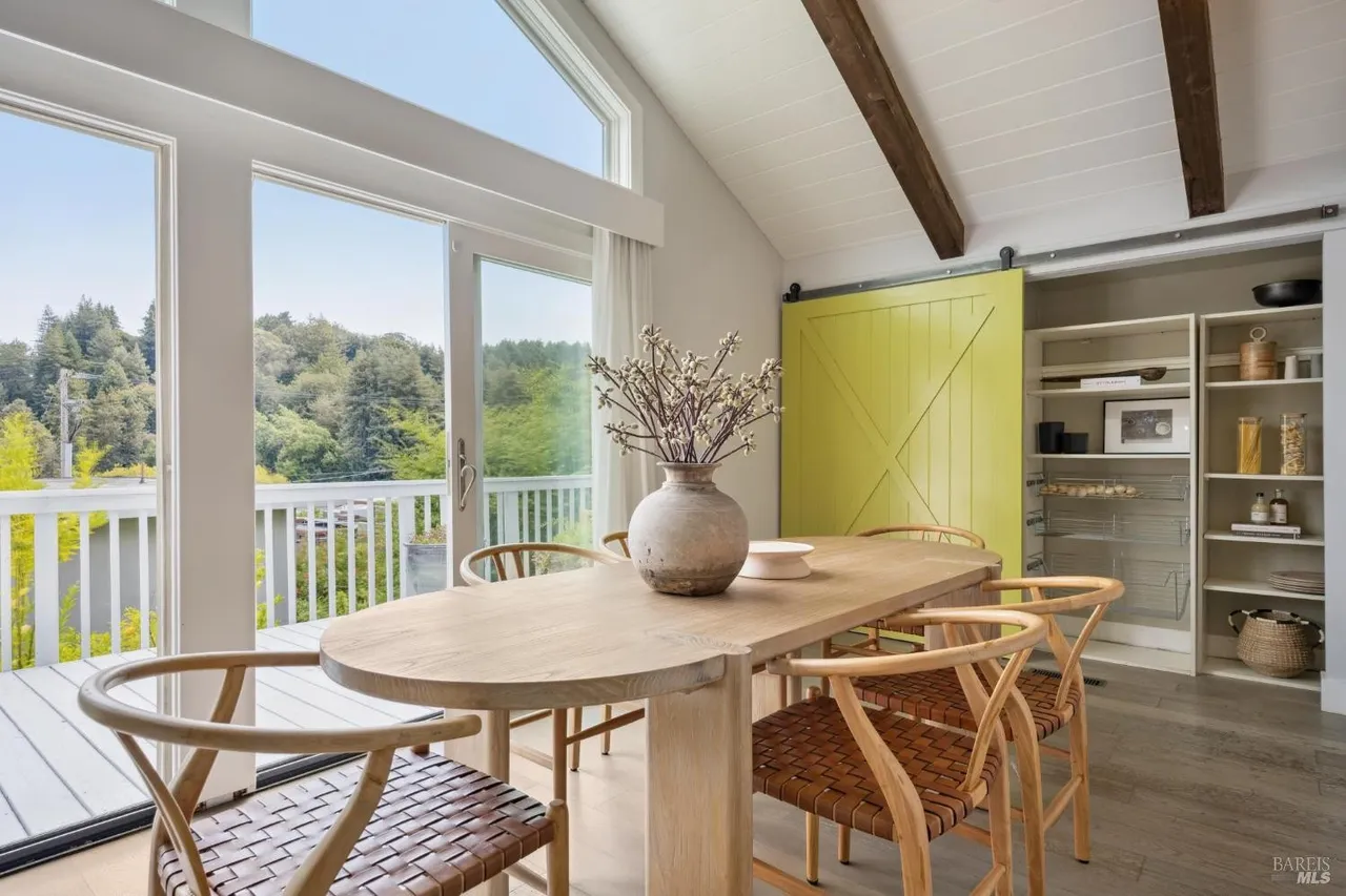 This dining room features a large, light wood oval table surrounded by woven chairs, creating a warm and inviting atmosphere. A sliding barn door in a vibrant yellow adds a pop of color, while large windows offer views of the outdoors, filling the space with natural light. The room is styled with a vase of branches as a centerpiece, enhancing the natural aesthetic.