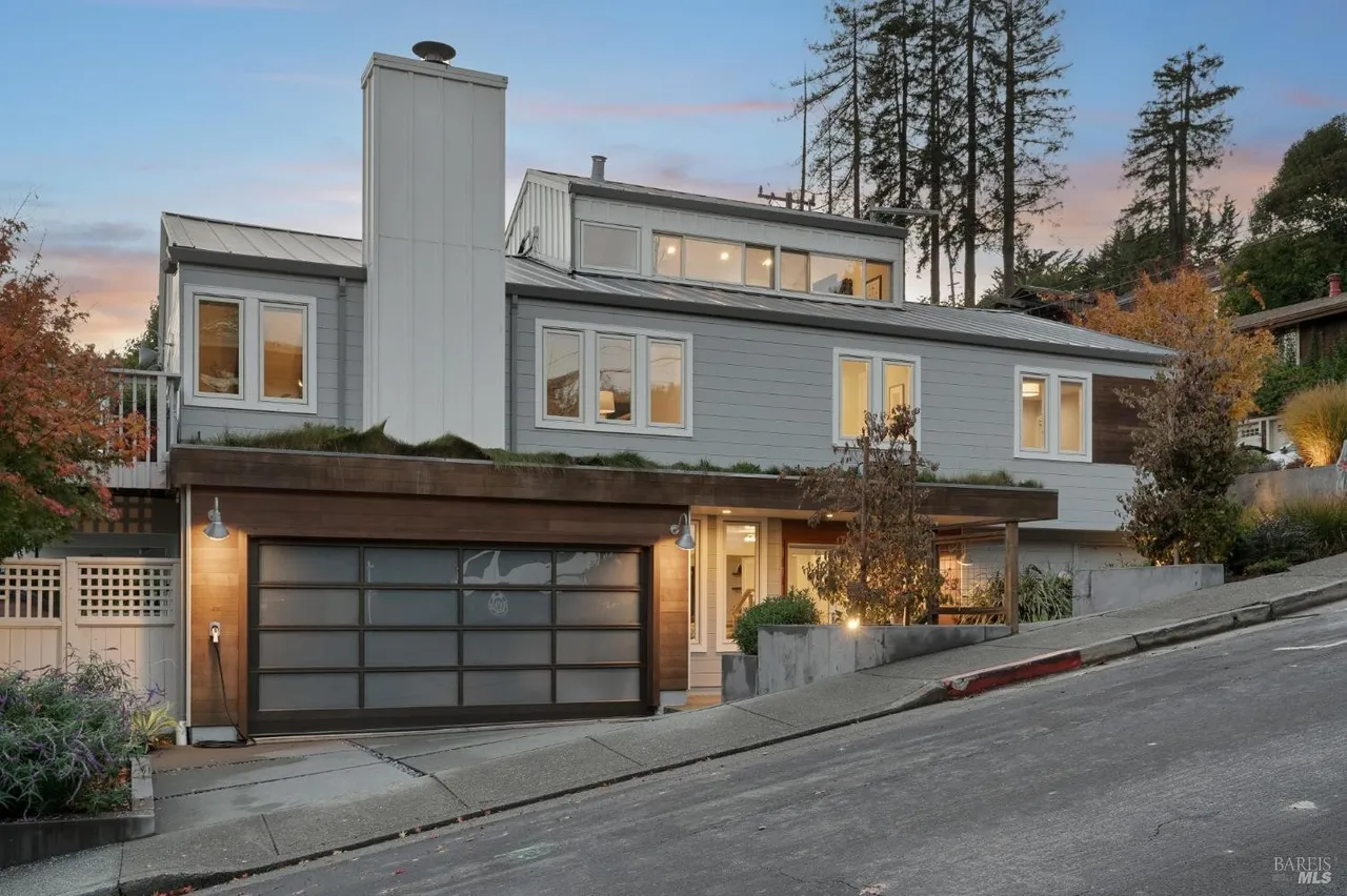 This is a front exterior view of a modern, multi-level home with a unique architectural design. The house features a gray exterior, a flat roof with a green roof element over the garage, and a prominent white chimney. A glass garage door and well-maintained landscaping add to the property's curb appeal, while the sloped driveway suggests a hillside location.