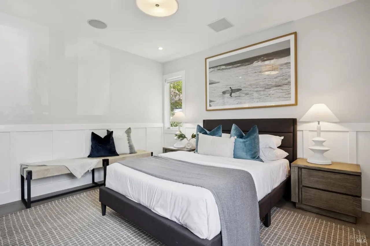 This is a bright and airy primary bedroom featuring a neutral color palette with pops of blue. The room includes a bed with a padded headboard, a nightstand with a lamp, and a bench at the foot of the bed. A large framed print hangs above the bed, and the room is well-lit with natural light from a window.