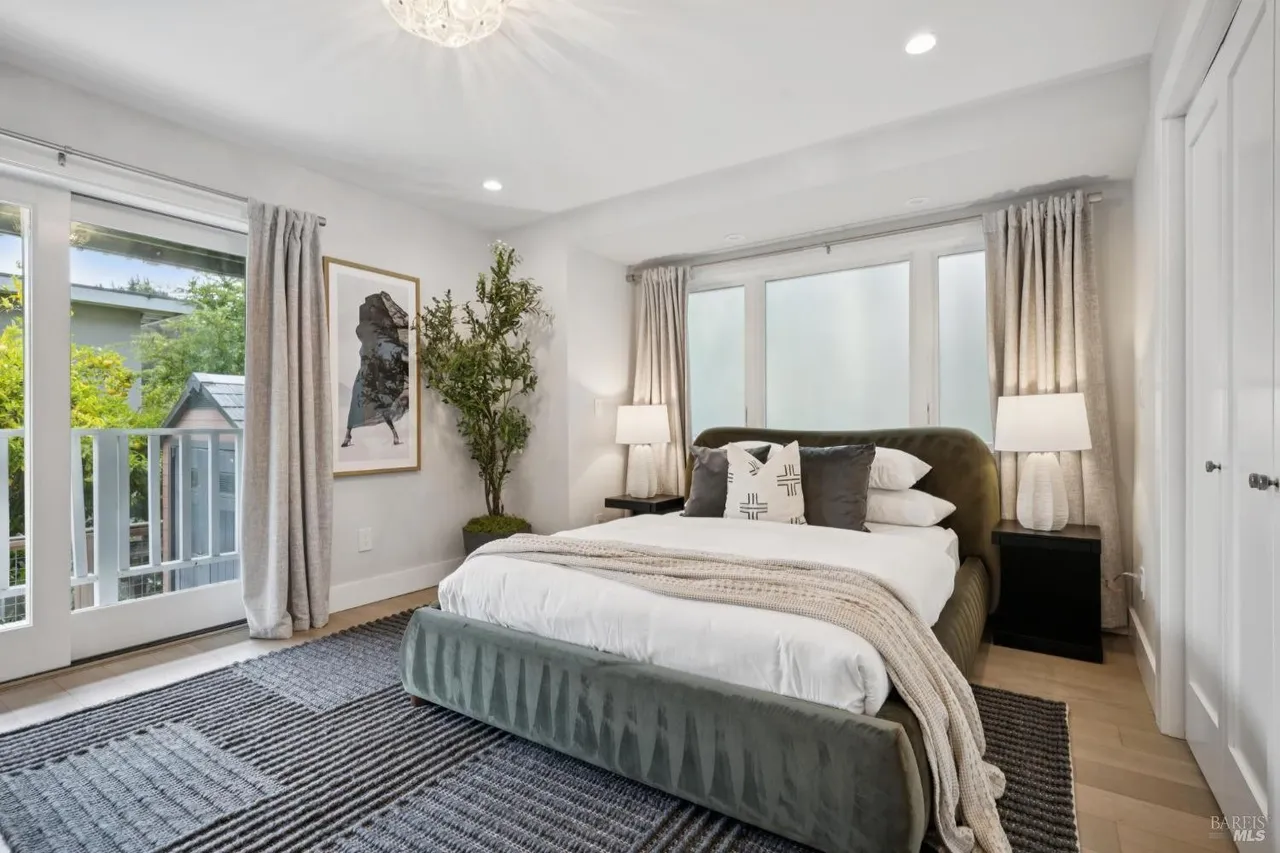 This is a primary bedroom featuring a large bed with a gray velvet frame, white linens, and decorative pillows. The room has neutral-toned walls, curtains, and a patterned rug. Natural light streams in through the windows, and there's a potted tree adding a touch of greenery. The overall impression is serene and stylish.