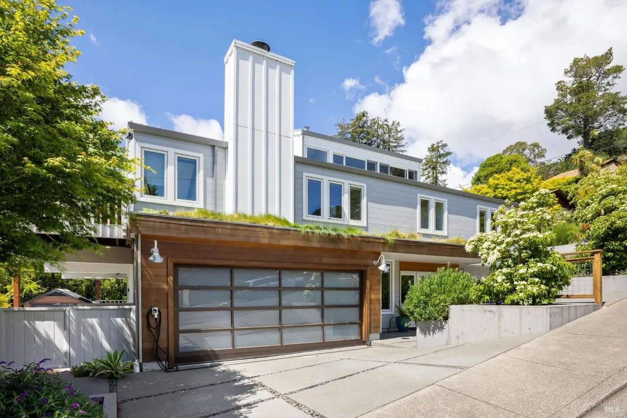 This is a front exterior view of a modern two-story home with a unique architectural design. The house features a large garage with a glass door, a green roof over the garage, and a tall, white chimney. The property includes a well-maintained yard and a concrete driveway, creating a welcoming and stylish curb appeal.