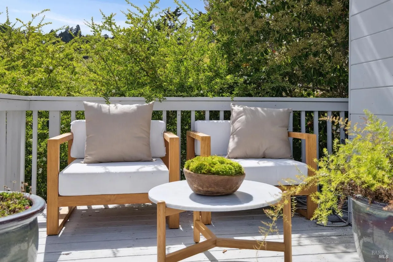 This image showcases a cozy outdoor seating area on a patio or deck. Two wooden-framed chairs with white cushions and neutral pillows are arranged around a small round table, creating an inviting space for relaxation. Lush greenery in the background and potted plants add to the serene atmosphere, enhancing the appeal of this outdoor living space.