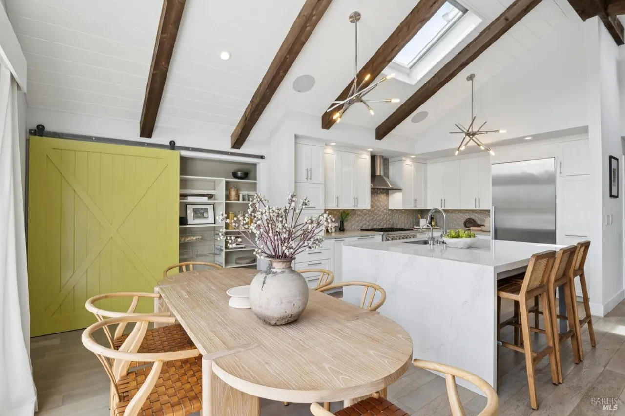 This interior shot showcases a bright, modern kitchen and dining area. The kitchen features white cabinetry, stainless steel appliances, and a large island with a marble countertop. A wooden dining table with woven chairs sits in the foreground, complemented by a unique yellow sliding barn door and exposed wooden beams on the ceiling, creating a warm and inviting atmosphere.