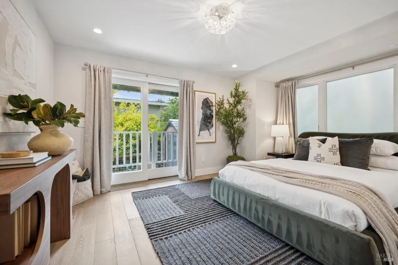 This is a bright and airy primary bedroom featuring a large bed with a gray velvet frame, white linens, and decorative pillows. The room has a neutral color palette with light hardwood floors and gray curtains framing a balcony door. A modern console table with a vase of greenery adds a touch of nature to the space.