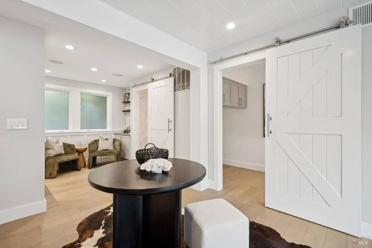 This interior shot showcases a stylish living room area with a modern farmhouse aesthetic. The room features light hardwood flooring, a dark round table with decorative items, and comfortable seating. A sliding barn door adds a rustic touch, while the overall design creates a cozy and inviting atmosphere.
