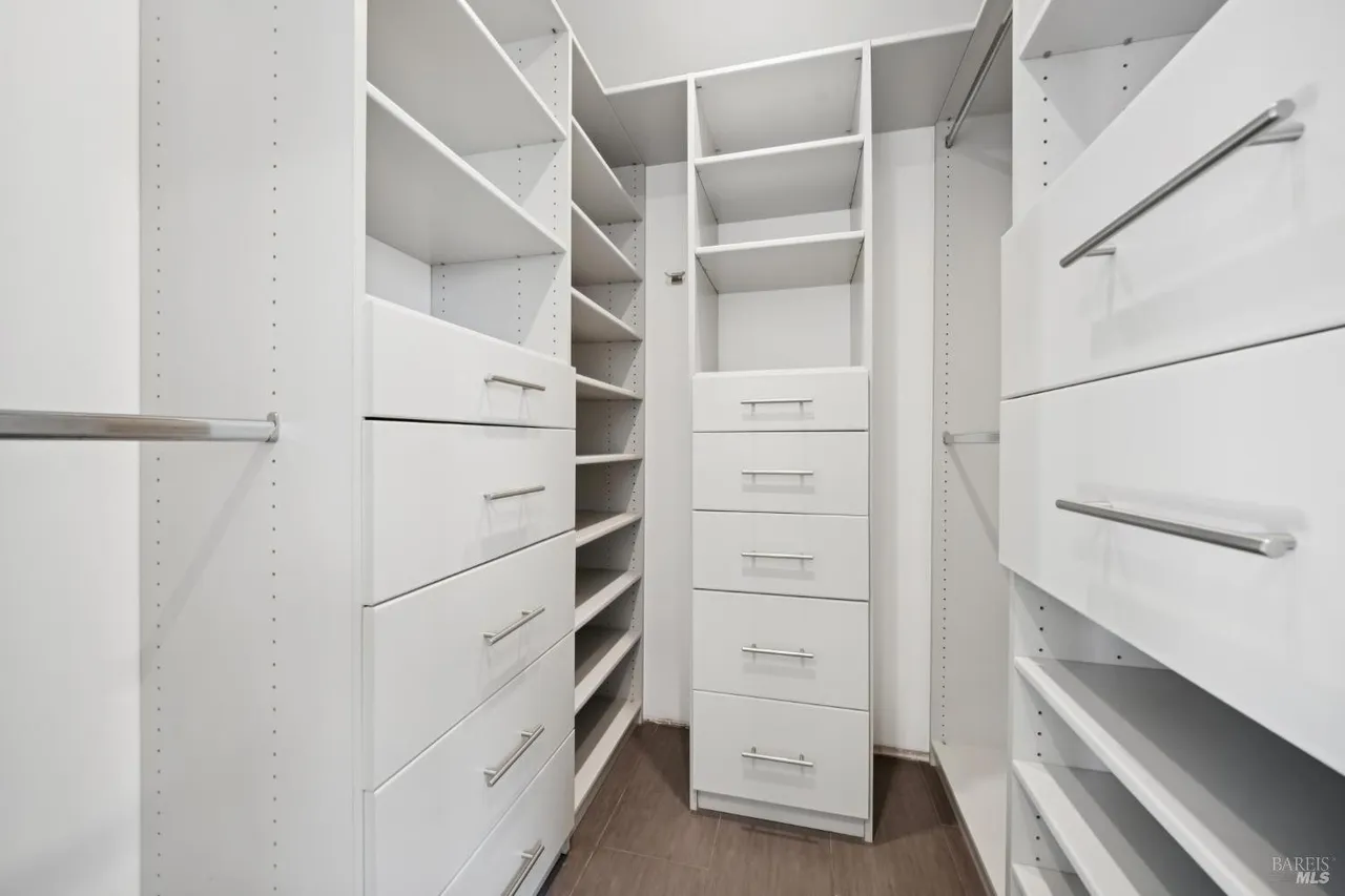 This is an interior shot of a well-organized walk-in closet. The closet features white shelving and drawers, providing ample storage space. The design is clean and modern, with silver hardware adding a touch of elegance.