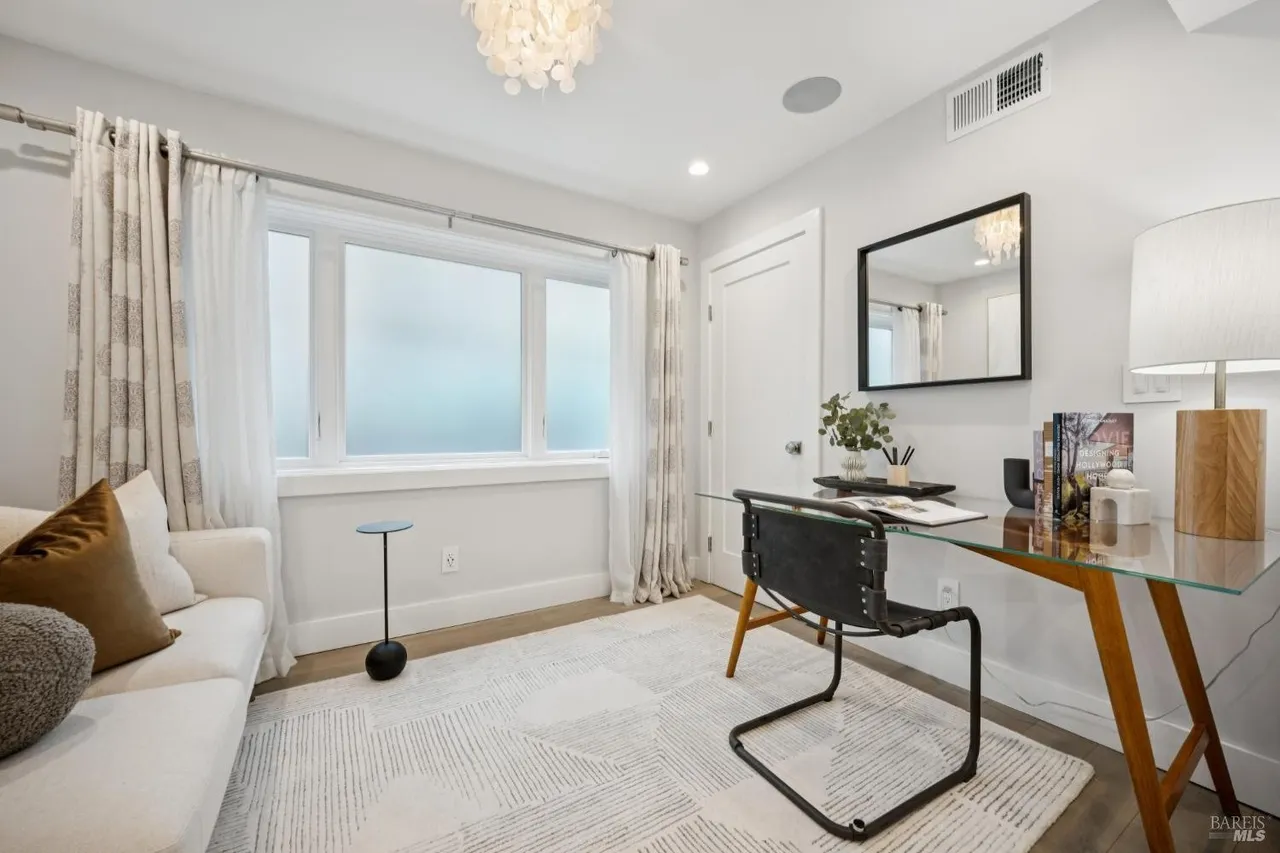 This is an interior shot of a home office featuring a glass-top desk with wooden legs and a black leather chair. The room is decorated in neutral tones with a white sofa, patterned curtains, and a modern chandelier. A window provides natural light, and a mirror hangs on the wall, adding depth to the space.