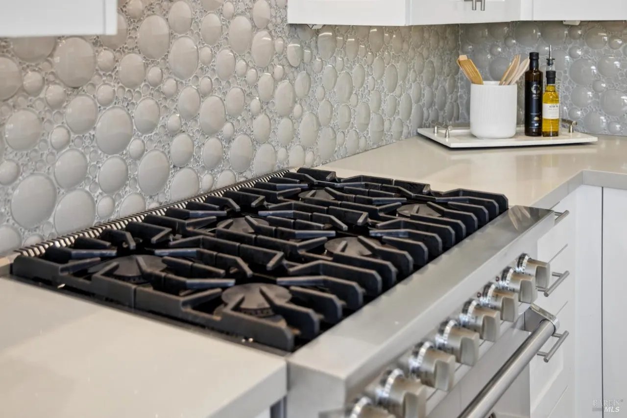 This is a close-up shot of a modern kitchen featuring a high-end gas range with a stainless steel finish and heavy-duty black grates. The countertop is a light, neutral color, and the backsplash is a unique, textured tile. The overall impression is one of luxury and functionality.