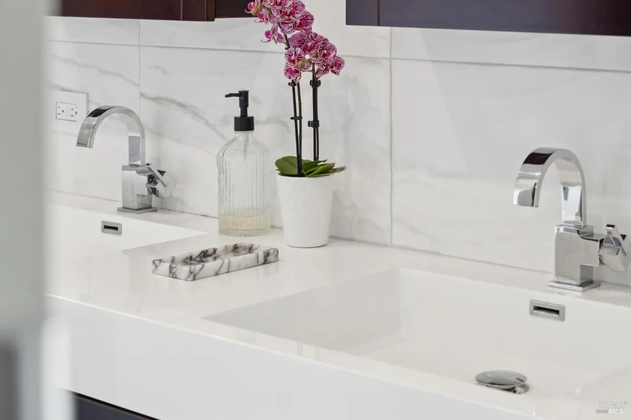 This is a detailed shot of a modern bathroom vanity featuring dual sinks, chrome faucets, and a white countertop. A decorative orchid and a soap dispenser add a touch of elegance. The overall impression is clean, bright, and stylish, highlighting the bathroom's contemporary design.