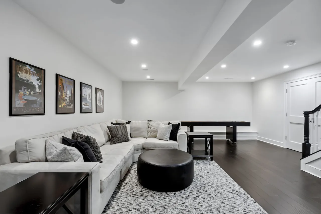 This is a spacious living room featuring a large sectional sofa with decorative pillows, a black ottoman, and a patterned area rug. The walls are painted white, and there are framed art pieces on the wall. The room has recessed lighting and dark hardwood floors, creating a modern and inviting atmosphere.