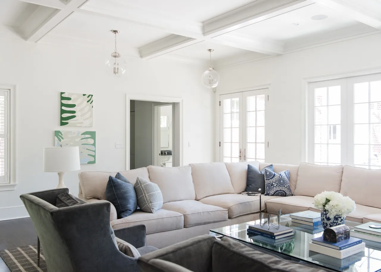 This is a bright and airy living room featuring a large, comfortable sectional sofa with blue and patterned throw pillows. The room is decorated in a neutral color palette with white walls and ceiling beams, complemented by modern artwork and glass pendant lighting. A glass coffee table sits in the center, adding to the room's sophisticated and inviting atmosphere.