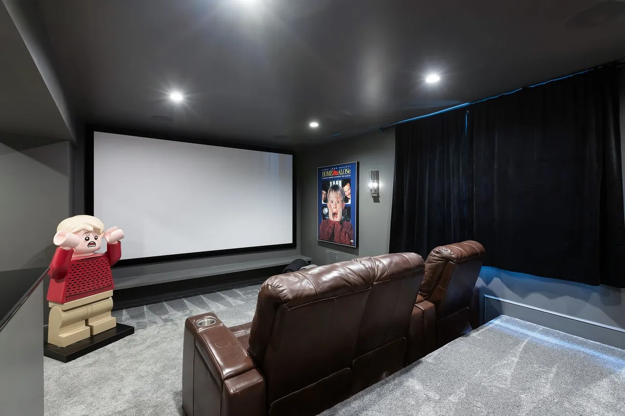 This is a home theater room featuring two brown leather reclining chairs facing a large projector screen. A 'Home Alone' movie poster hangs on the wall, and a large Lego figure stands to the side of the screen. The room has dark walls and ceiling, creating a cinematic atmosphere.
