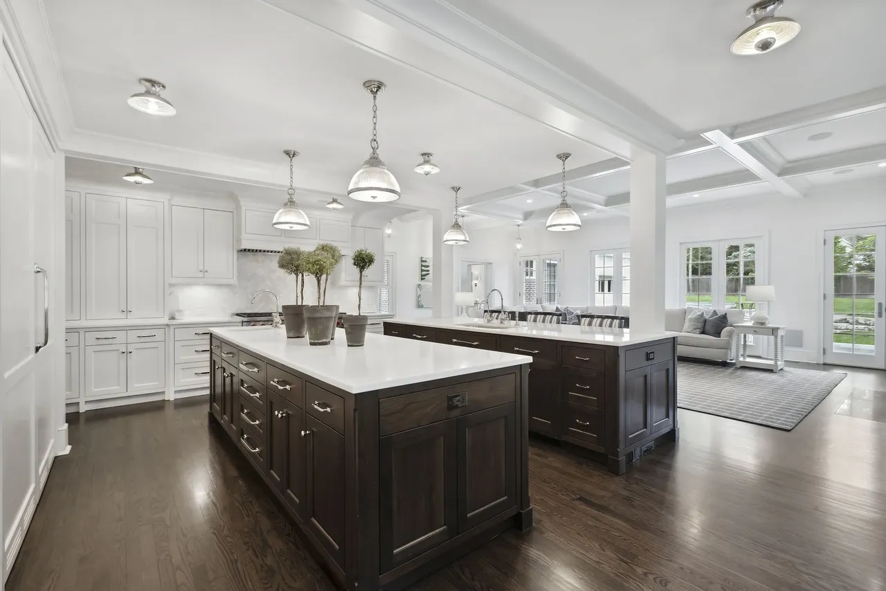 This is a bright and spacious kitchen featuring two large islands with dark wood cabinetry and white countertops. The kitchen is equipped with white cabinets and stainless steel appliances. The open floor plan leads into a living area with large windows, creating a seamless transition between spaces.
