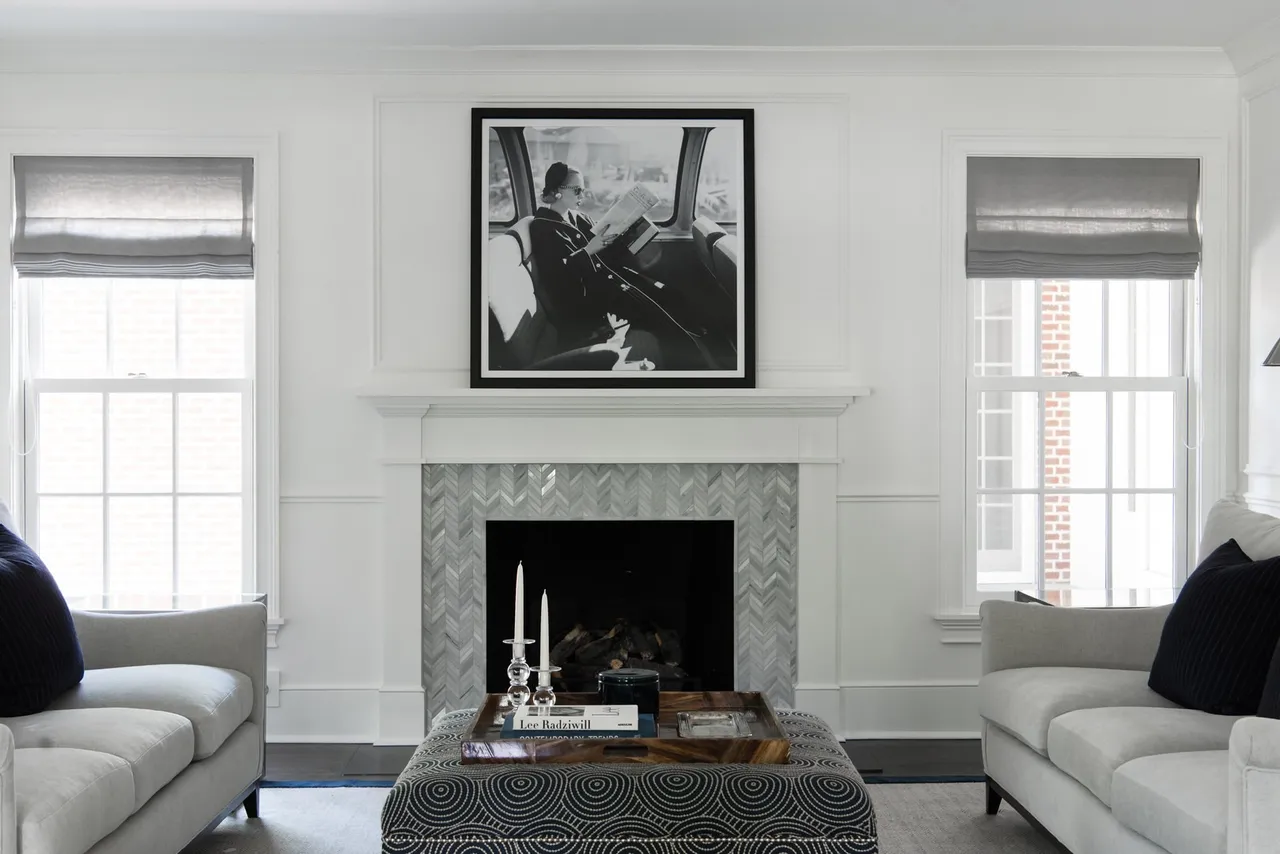 This living room features a classic and elegant design with a focus on symmetry. Two large windows flank a fireplace with a herringbone tile surround and a black and white photograph above the mantel. The room is furnished with light gray sofas and a patterned ottoman, creating a sophisticated and inviting atmosphere.