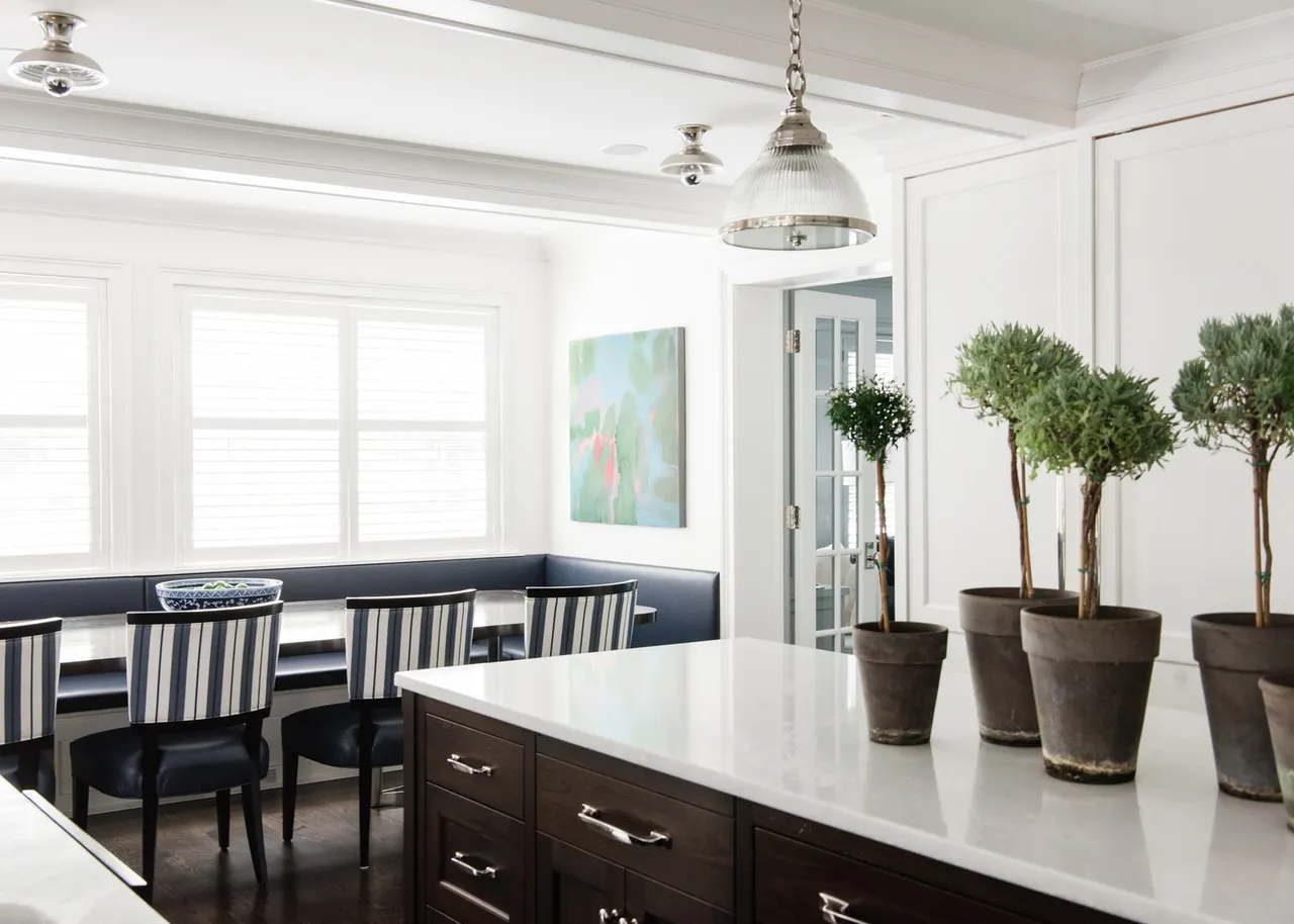 This is a bright and airy kitchen featuring a breakfast nook with a built-in bench and striped chairs. The kitchen island has a white countertop and dark wood cabinetry, topped with potted greenery. A pendant light hangs above the island, and a painting adorns the wall, adding a touch of color to the otherwise neutral palette.