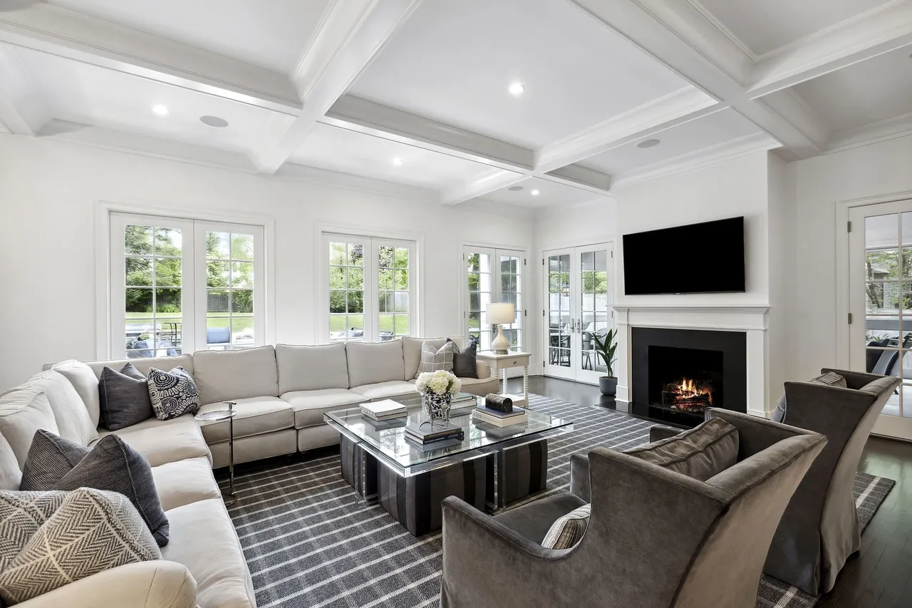 This is a bright and spacious living room featuring a large sectional sofa, a glass-topped coffee table, and two armchairs. A fireplace with a mounted television serves as a focal point, and multiple windows and French doors provide ample natural light and views of the outdoors. The room is decorated in a neutral color palette with a striped rug, creating a sophisticated and inviting atmosphere.