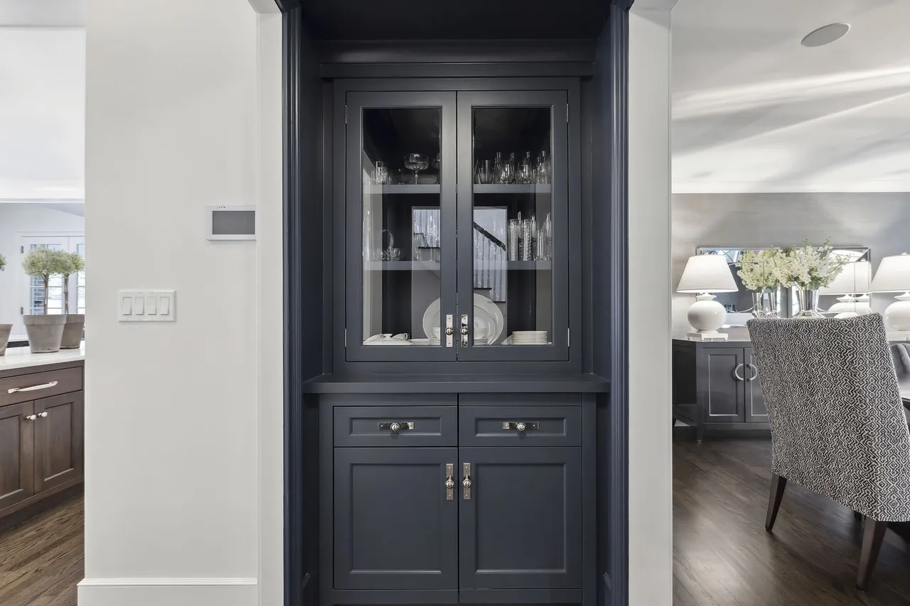 This image showcases a built-in china cabinet in a dining area, featuring glass-fronted upper cabinets displaying glassware and plates, and closed lower cabinets with elegant hardware. The dark blue-gray cabinetry provides a sophisticated contrast to the white walls, while the glimpse into the dining area reveals a stylish chair and sideboard, suggesting a well-appointed and inviting space.