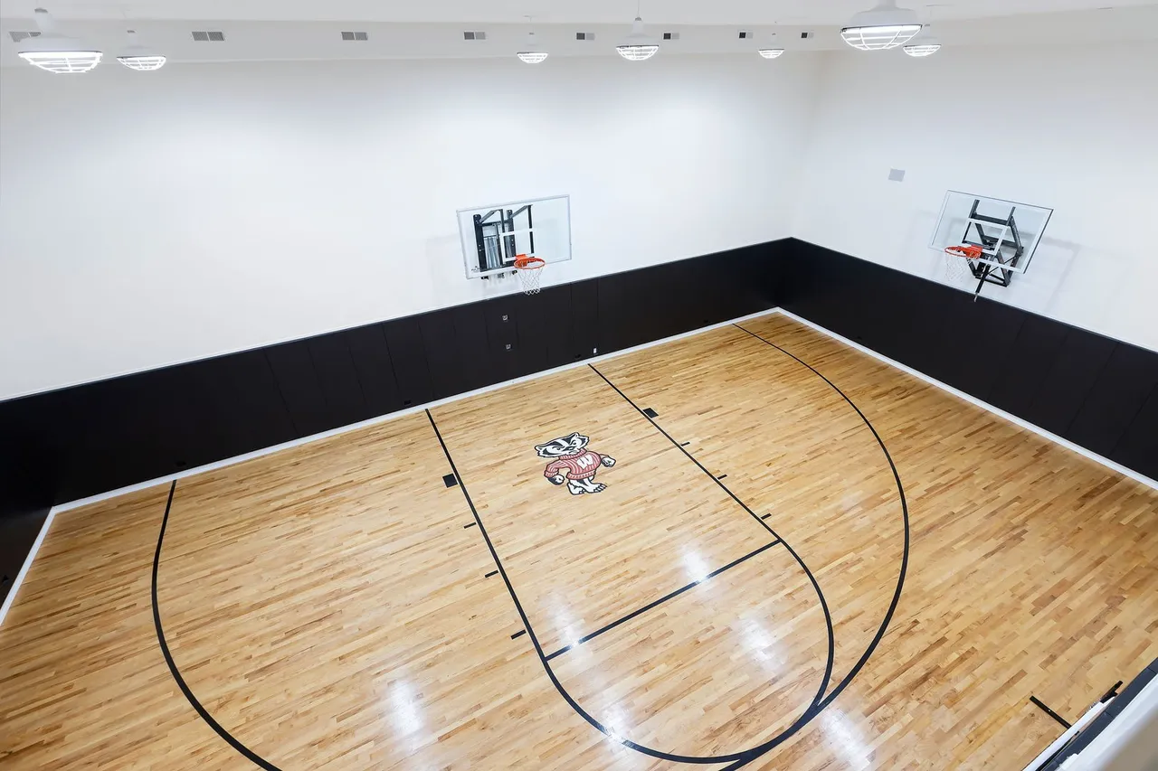 This is an indoor basketball court with a wooden floor featuring the Wisconsin Badgers logo at center court. The walls are white with a black padded section along the lower portion, and there are two basketball hoops mounted on the walls. The space is well-lit with multiple ceiling lights, creating a bright and inviting atmosphere for athletic activities.