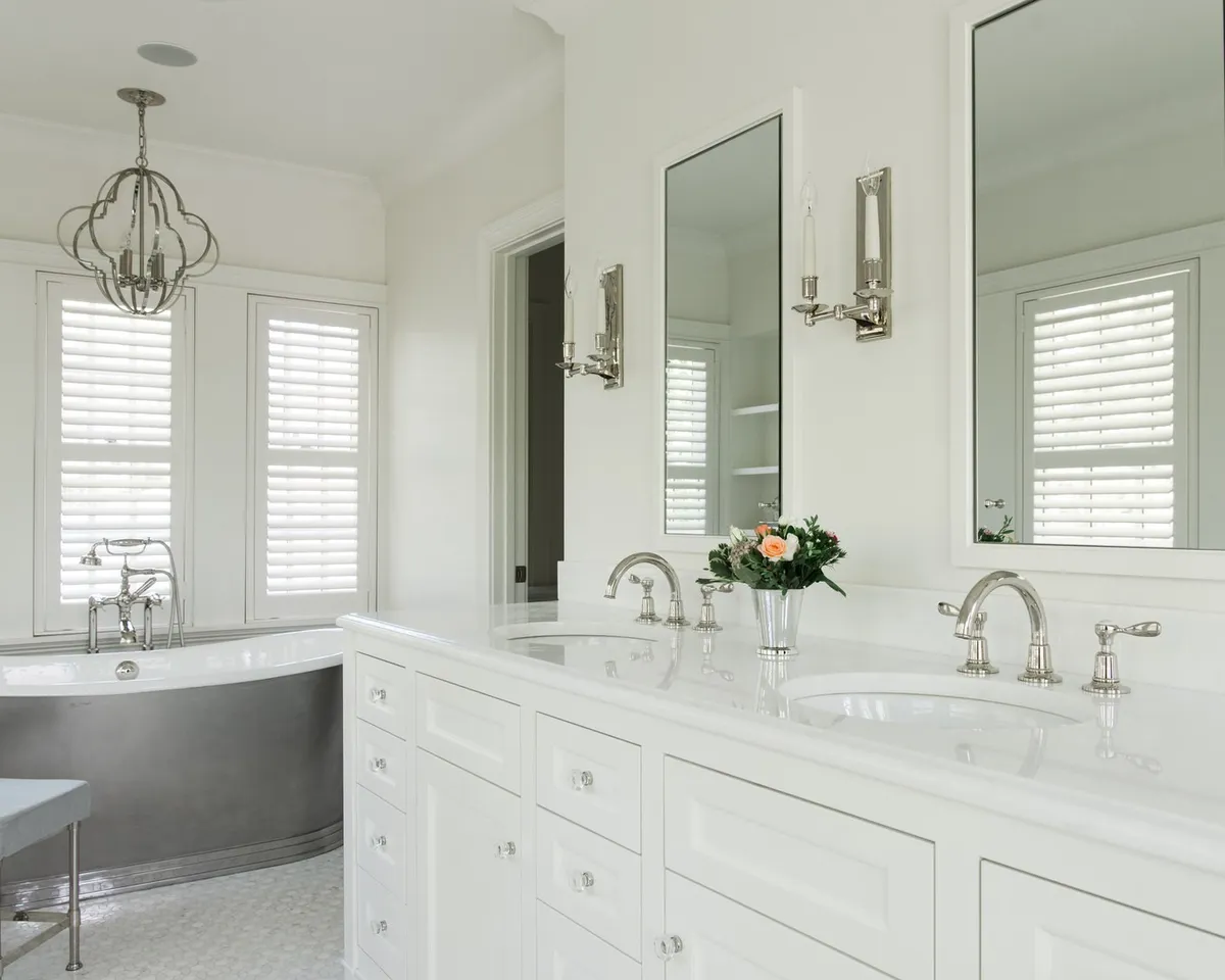 This is a bright and elegant primary bathroom featuring a double vanity with white cabinetry and marble countertops. Two framed mirrors hang above the sinks, flanked by sconces. A freestanding bathtub sits beneath shuttered windows, adding a touch of luxury to the space.