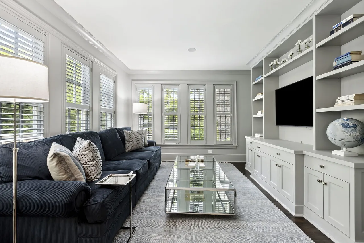 This is an interior shot of a living room featuring a large, comfortable navy blue corduroy sofa with decorative pillows. A glass-topped coffee table sits in the center of the room on a gray rug, and a built-in shelving unit with a television provides storage and entertainment. Natural light floods the room through multiple windows with white shutters, creating a bright and inviting atmosphere.
