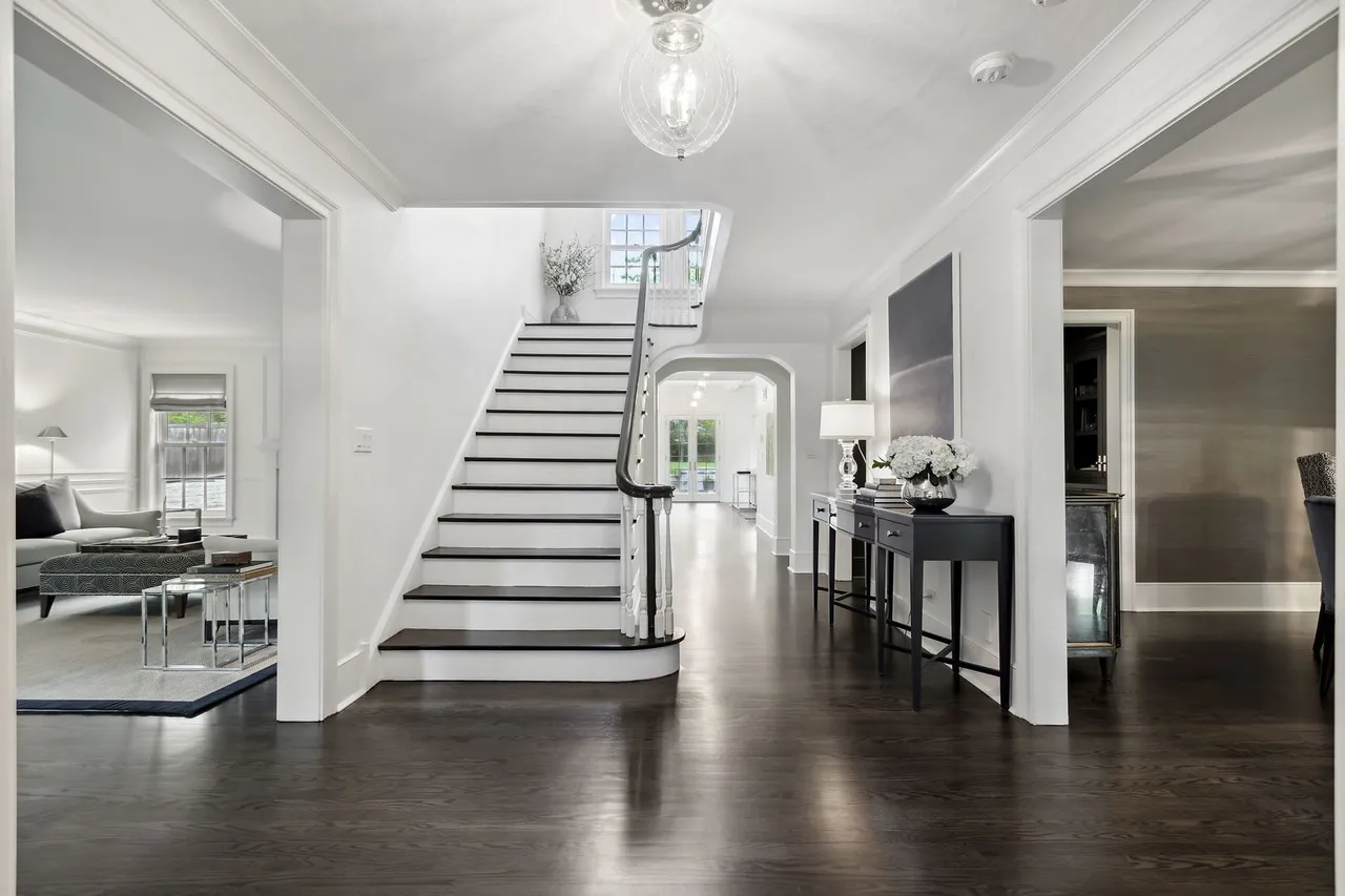 This is an interior shot of a grand foyer featuring a staircase with black treads and a white riser. The dark hardwood floors reflect light, adding warmth to the space. Two console tables with lamps and floral arrangements flank the hallway, leading to other rooms in the house. The overall impression is elegant and inviting.