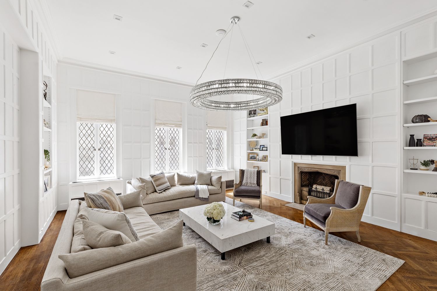 This is a bright and elegant living room featuring a neutral color palette with white walls and light-colored furniture. The room is well-lit with natural light from the windows and a large, decorative chandelier. A fireplace with a mounted television above it serves as a focal point, flanked by built-in bookshelves, creating a sophisticated and inviting atmosphere.