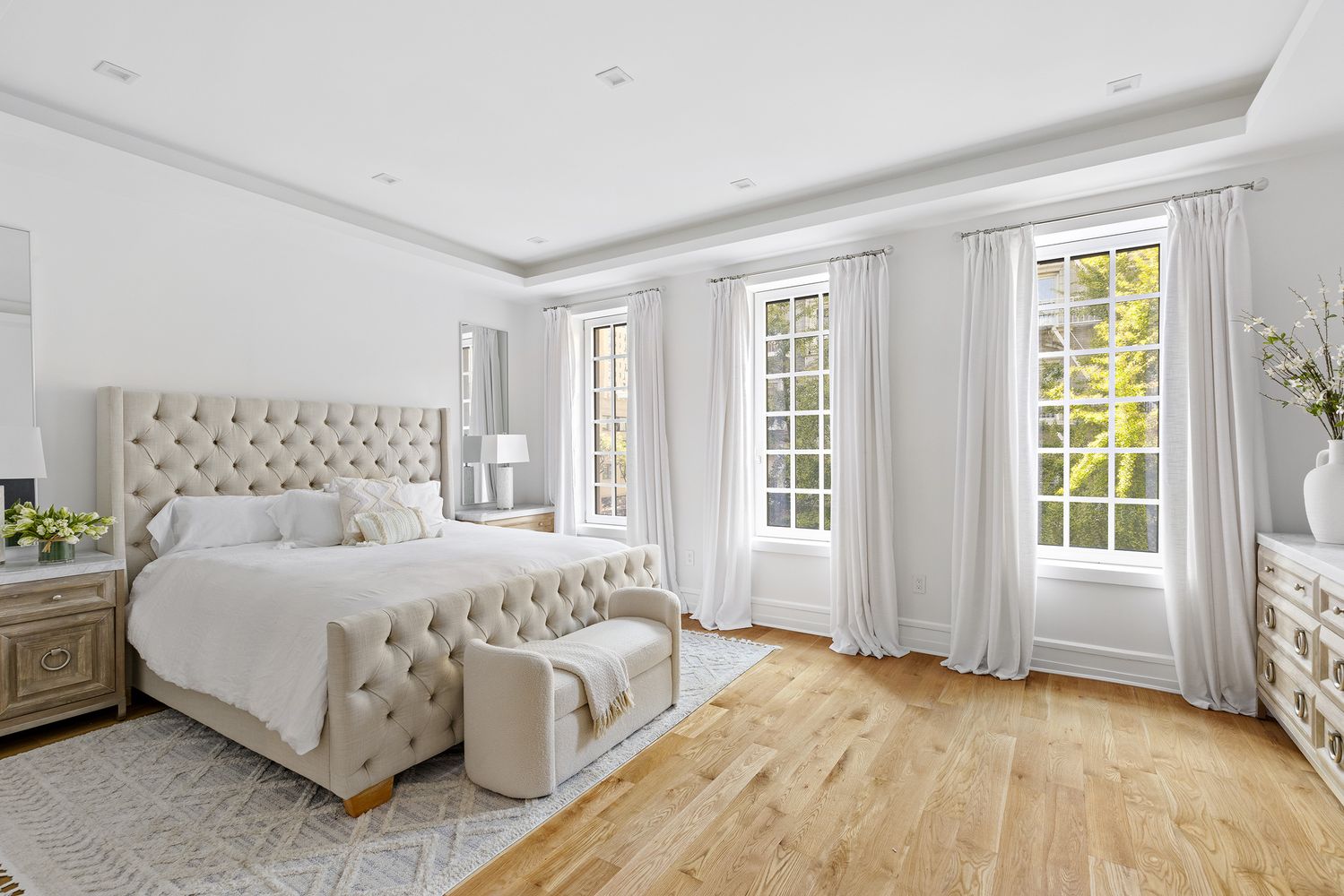 This is a bright and spacious primary bedroom featuring a large, tufted headboard bed with white linens and a matching bench. The room is illuminated by natural light streaming through three large windows dressed with white curtains. Light hardwood floors and neutral-toned furniture create a serene and luxurious atmosphere.