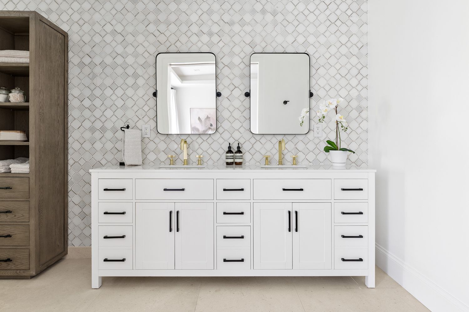 This is a well-lit primary bathroom featuring a double vanity with a white cabinet and black hardware. The backsplash is a patterned tile, and there are two rectangular mirrors above the sinks. A tall linen cabinet is on the left, and the overall style is modern and elegant.