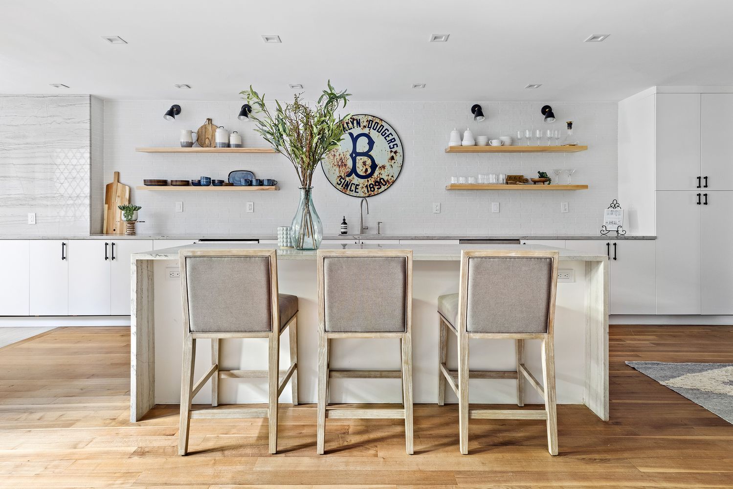 This is a bright and modern kitchen featuring a large island with three bar stools, white cabinetry, and open shelving. The backsplash is white subway tile, and there is a decorative Brooklyn Dodgers sign above the sink. The hardwood floors add warmth to the space, creating an inviting atmosphere.