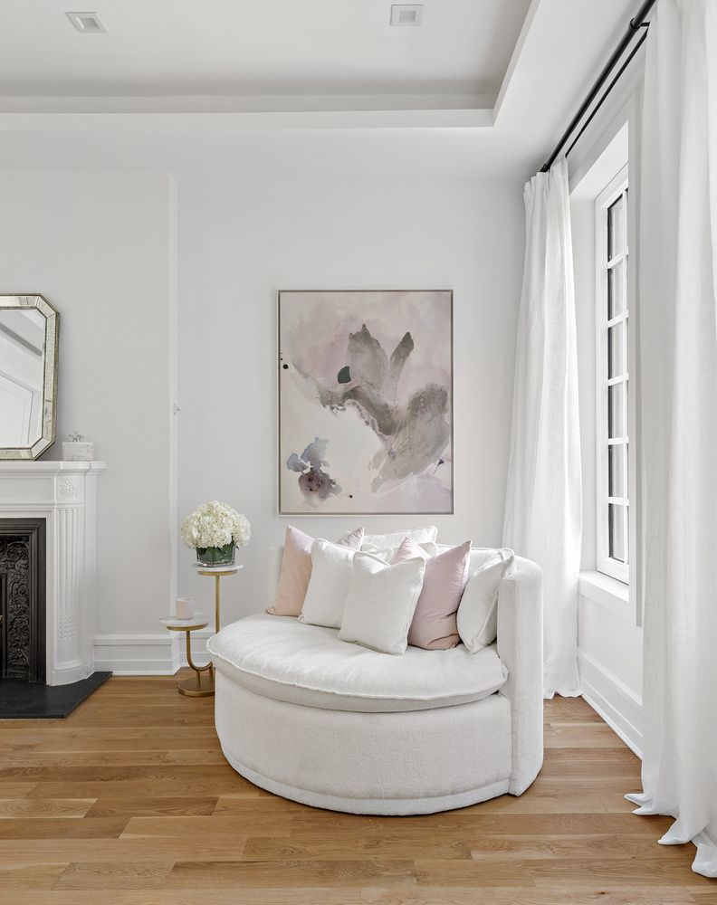 This is an interior shot of a living room featuring a round, white, plush sofa with decorative pillows, positioned in front of a large abstract painting. The room is well-lit with natural light streaming through a window draped with white curtains. The hardwood floors and fireplace add warmth and character to the space, creating an inviting and elegant atmosphere.