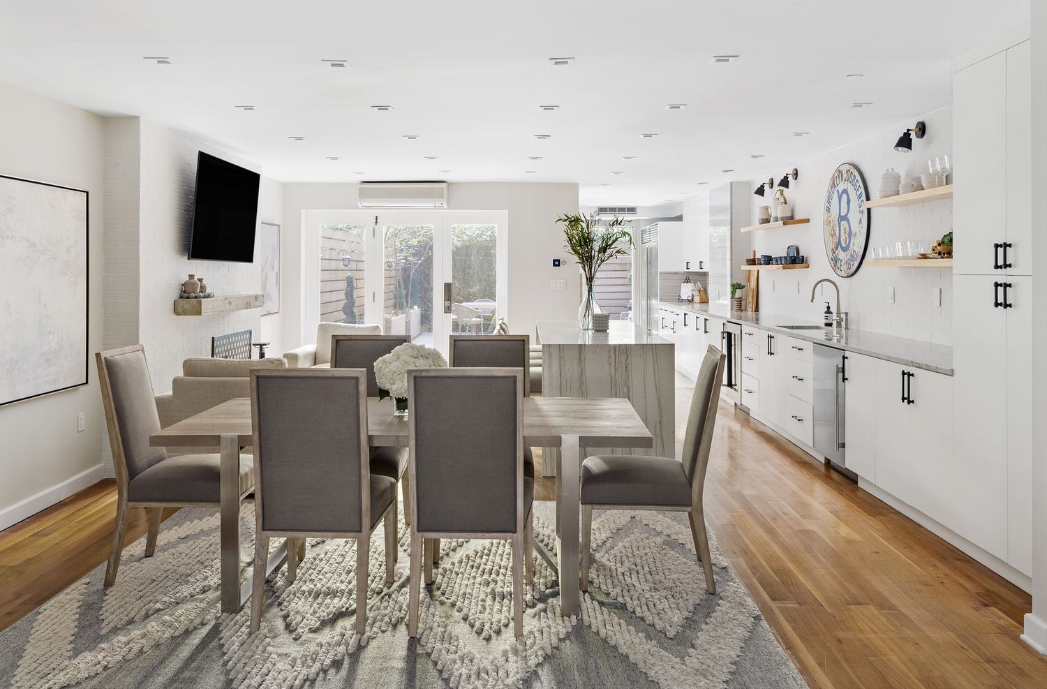 This is an interior shot of a dining room that seamlessly transitions into a modern kitchen. The dining area features a wooden table with six upholstered chairs, set upon a patterned rug. The kitchen boasts white cabinetry, stainless steel appliances, and a marble-topped island, creating a bright and airy atmosphere.