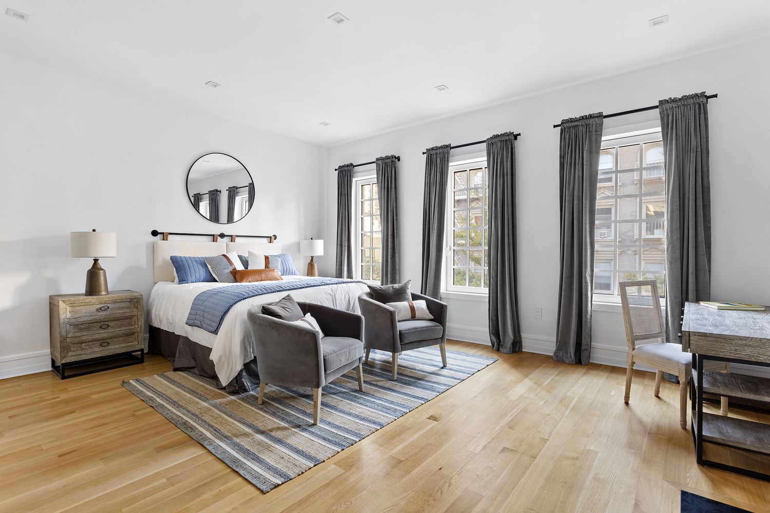 This is a bright and airy primary bedroom featuring a king-size bed with blue and white bedding, flanked by matching nightstands and lamps. Two gray armchairs sit on a striped rug in front of the bed, adding a cozy seating area. Three large windows with gray curtains provide ample natural light, highlighting the hardwood floors and white walls.