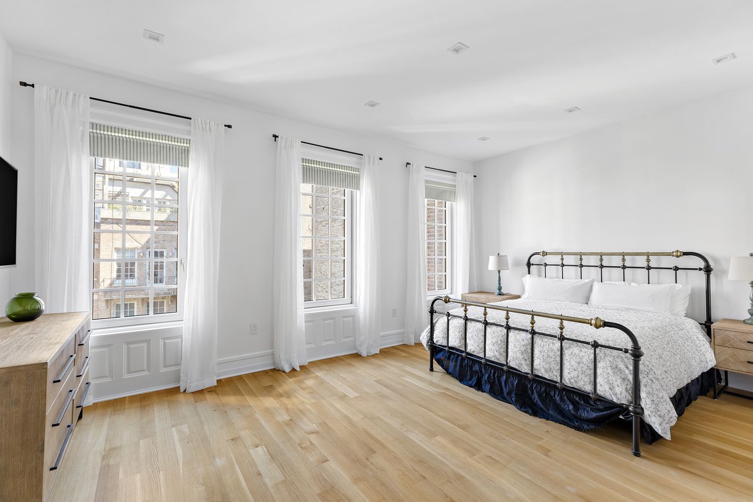 This is a bright and airy primary bedroom featuring hardwood floors, white walls, and three large windows dressed with sheer white curtains. A black metal bed frame with brass accents is the focal point, complemented by a dark blue bed skirt and white bedding. The room exudes a clean and serene atmosphere, perfect for relaxation.