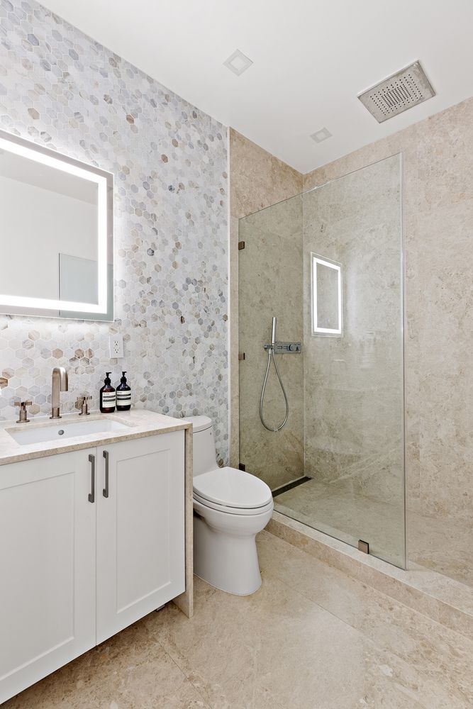 This is a well-lit bathroom featuring a modern design with a white vanity, a light-up mirror, and a toilet. The shower is enclosed with glass and has a rainfall showerhead. The walls are tiled with a mosaic pattern, and the floor is tiled with a light-colored stone, creating a clean and luxurious feel.