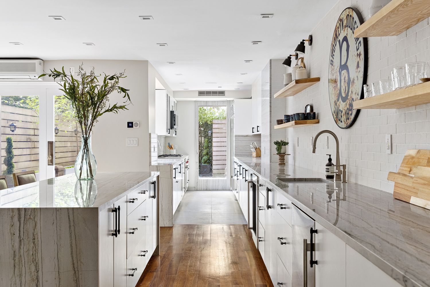 This is a bright and modern kitchen featuring white cabinetry, marble countertops, and stainless steel appliances. The kitchen has an open layout with a large island and a view to the backyard through a glass door. The hardwood floors add warmth to the space, and the overall design is clean and elegant.