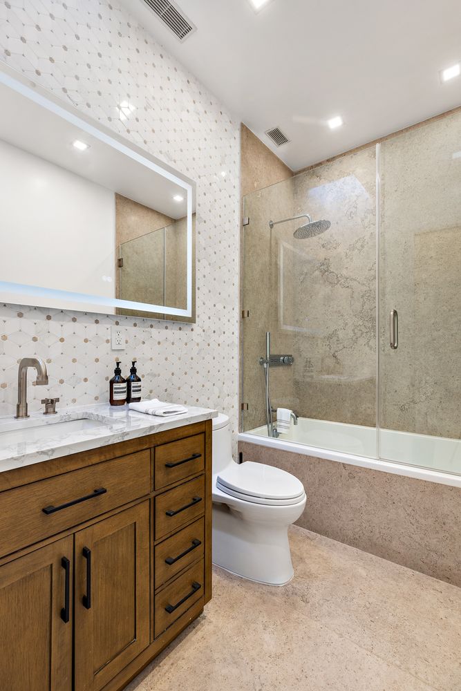 This is a well-lit bathroom featuring a wooden vanity with a marble countertop, a modern faucet, and a large, illuminated mirror. The walls are decorated with patterned tiles, and a glass-enclosed bathtub/shower combination is present. The overall impression is clean, stylish, and functional.