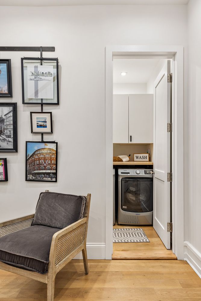 This image showcases a well-organized laundry area, accessible through a doorway. The laundry room features a modern washing machine, white cabinetry above, and a wooden shelf with a "WASH" sign. A gray and white patterned rug adds a touch of style to the space, while a comfortable chair sits just outside the laundry room.