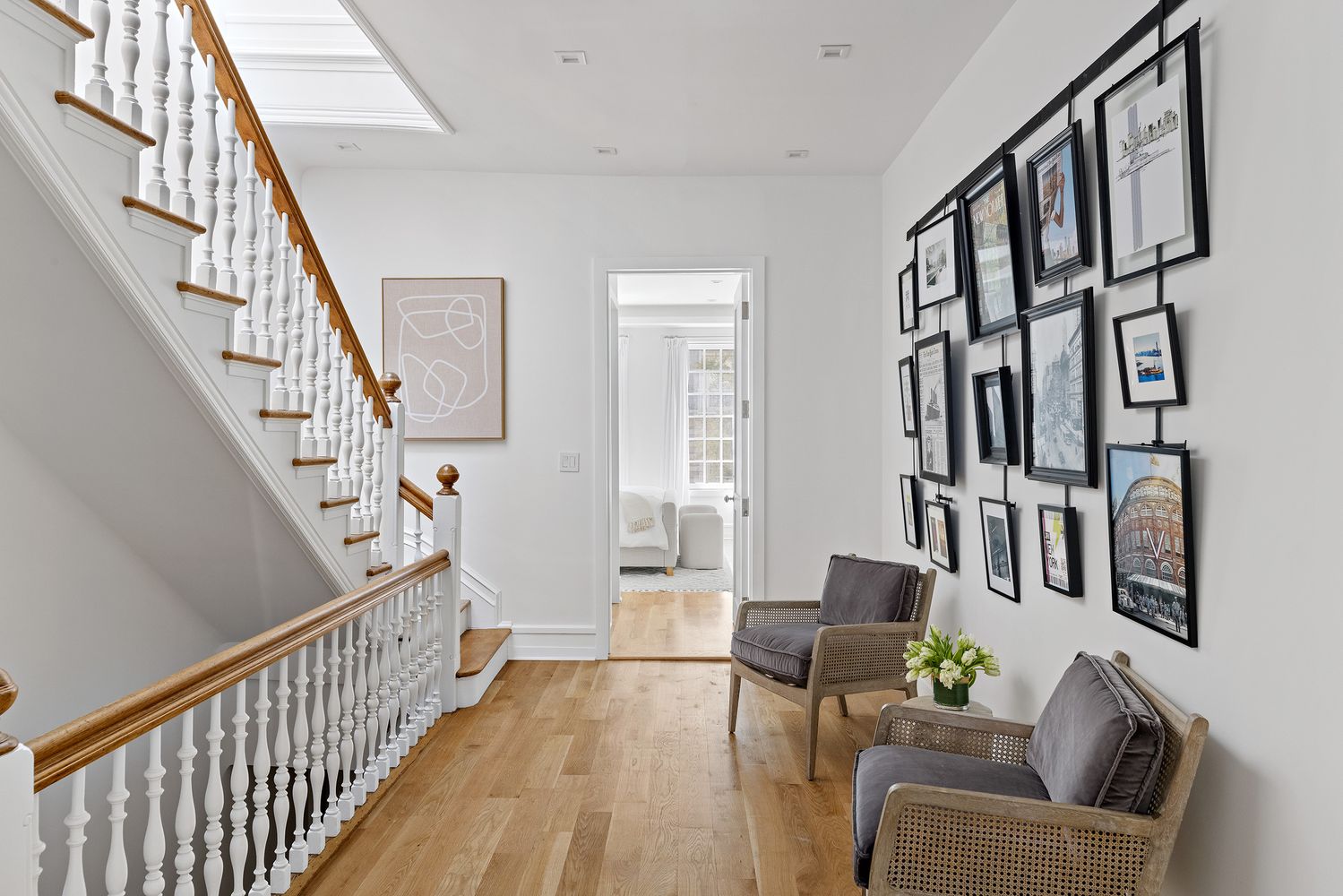 This interior shot showcases a bright and inviting hallway with a staircase. The hallway features hardwood floors, white walls adorned with a gallery wall of framed pictures, and two armchairs with gray cushions. The staircase has white spindles and a wooden handrail, leading to an upper level, while an open doorway provides a glimpse into another room, creating a sense of depth and flow.