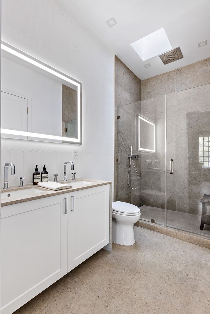 This is a well-lit primary bathroom featuring a double vanity with white cabinets and a light-colored countertop. A large, illuminated mirror hangs above the vanity. The shower is enclosed with glass and has neutral-toned tile, and a toilet is positioned next to the shower. The floor is a speckled beige tile, creating a clean and modern aesthetic.