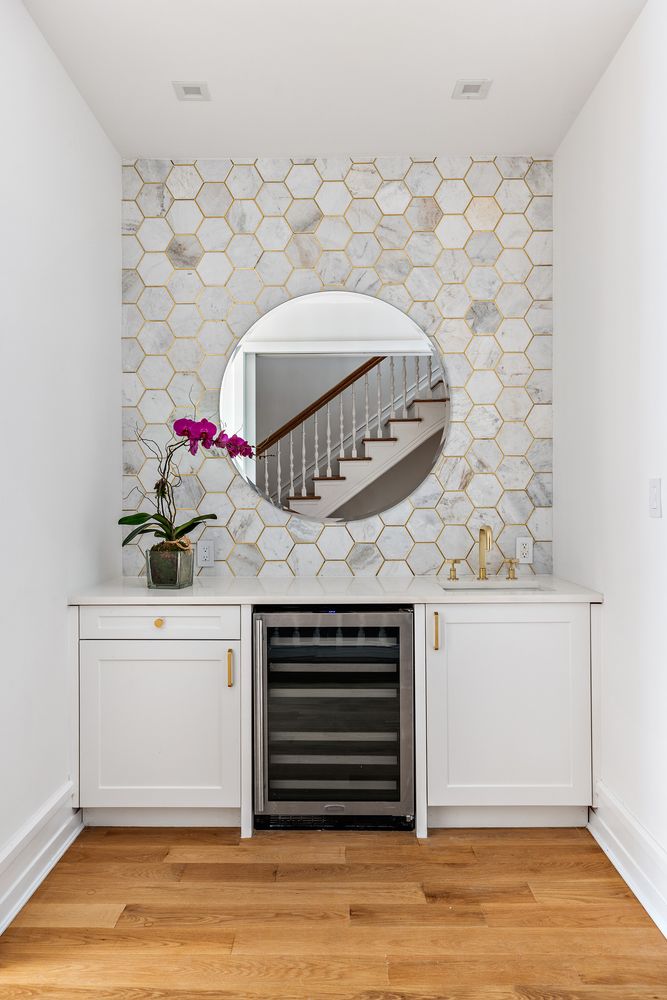 This interior shot showcases a stylish hallway area featuring a built-in bar with white cabinetry, gold hardware, and a wine cooler. A round mirror reflects the staircase, adding depth and visual interest to the space, while the hexagon tile backsplash with gold accents complements the hardwood flooring, creating a sophisticated and inviting atmosphere.