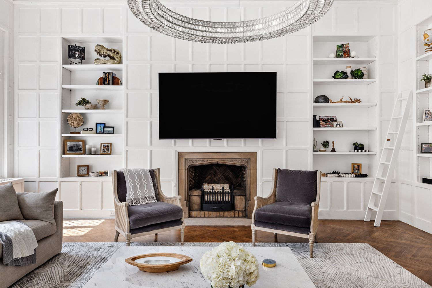 This is an interior shot of a living room featuring a fireplace with a large television mounted above it. The room is decorated with built-in bookshelves on either side of the fireplace, filled with decorative items. Two armchairs face the fireplace, and a coffee table with floral arrangements sits in the center of the room, creating a cozy and elegant atmosphere.