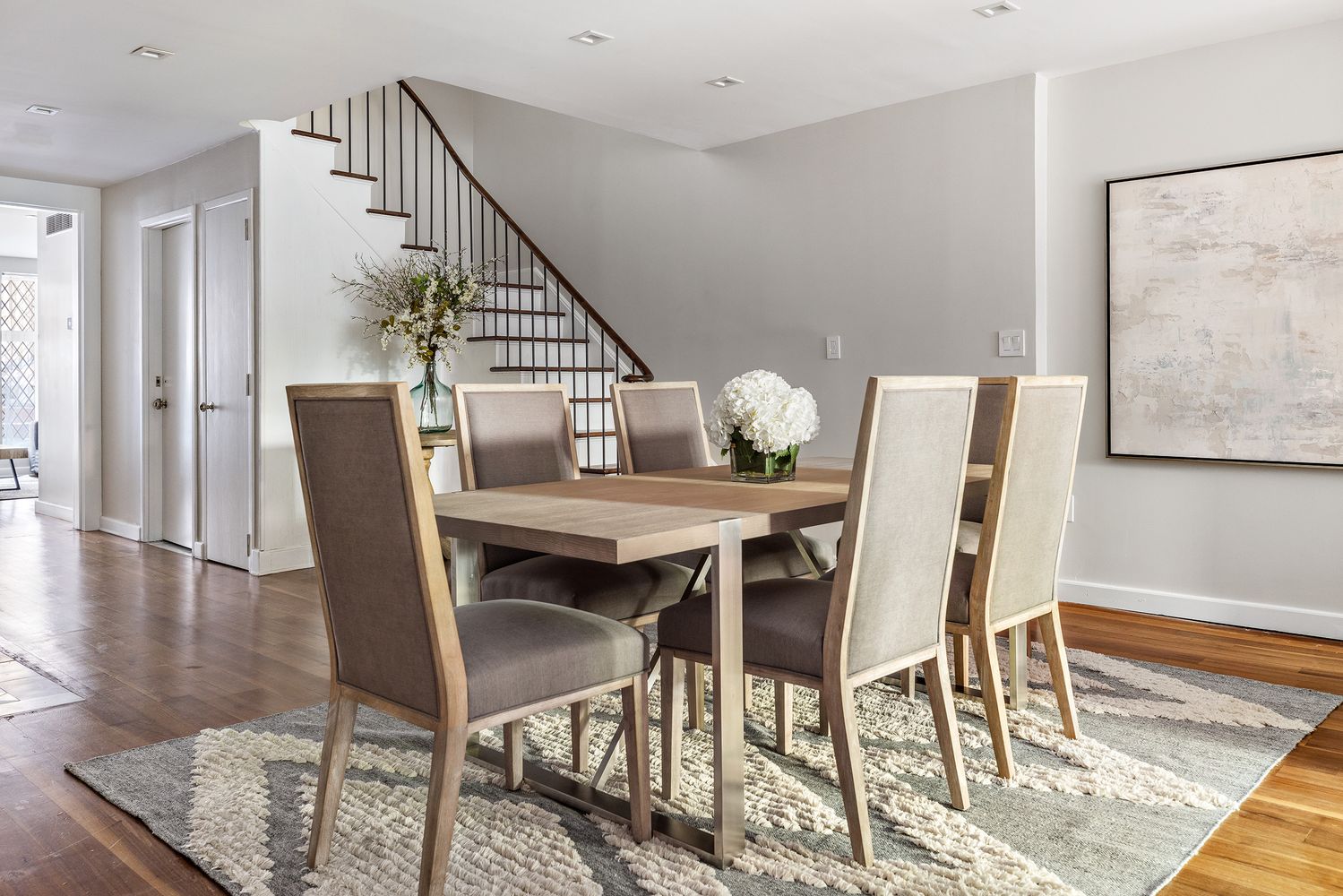 This is an interior shot of a dining room featuring a wooden dining table with six upholstered chairs. A patterned rug lies beneath the table, and a staircase is visible in the background. The room is well-lit and decorated with floral arrangements, creating a warm and inviting atmosphere.